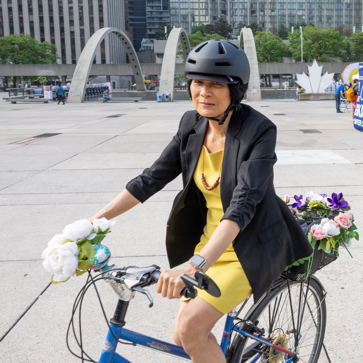 Olivia Chow riding a bicycle with a basket of flowers across Nathan Phillips Square